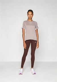 Naine seisab seljas heledat mauve värvi lühikeste varrukatega spordipead, tumedat pruunist leggingit logoga ja valgeid jooksujalanõusid roosa põhjaga.