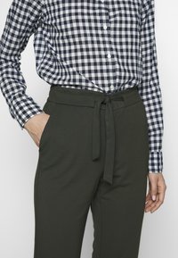 Person, die ein blau-weiß kariertes Hemd in eine hochgeschnittene dunkelgrüne Hose mit einem gebundenen Stoffgürtel gesteckt hat, mit der Hand in der Tasche.