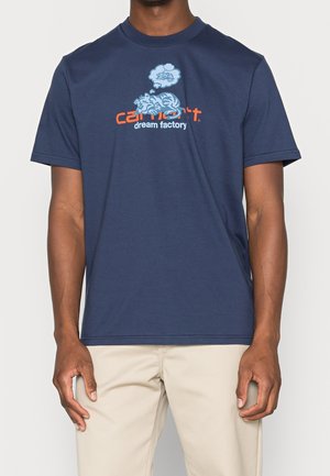 Mężczyzna w granatowej koszulce z nadrukiem śpiącego tygrysa i tekstem "Carhartt dream factory" na piersi.