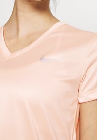 Vaaleanpunainen, lyhythihainen urheilu T-paita, jossa on V-pääntie ja Nike-logo hillityssä sävyssä. Kangas näyttää sileältä ja hengittävältä.
