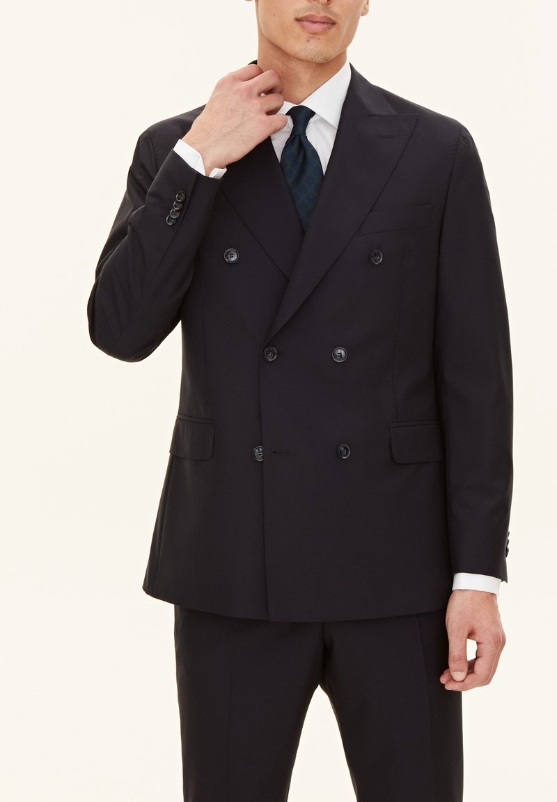 Granatowy garnitur dwurzędowy z klapami w kształcie notcha, dwoma przednimi kieszeniami i guzikami ozdobnymi. Nosi się go na białej koszuli i zielonym krawacie w wzory.