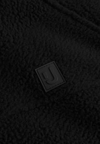 Tessuto in pile nero con una superficie texturizzata, che presenta un'etichetta quadrata nera in gomma con un logo stilizzato "J" nell'angolo.