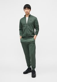 Αθλητικό φόρμα σε σκούρο πράσινο ύφασμα, που περιλαμβάνει ζακέτα με φερμουάρ και λογότυπο, ταιριαστό παντελόνι φόρμας με τσέπες, και μαύρα αθλητικά παπούτσια.