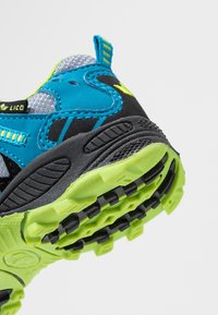 Zapatilla deportiva con parte superior azul y negra, suela externa de goma texturizada con acentos en verde brillante y asa para ponerse fácilmente.