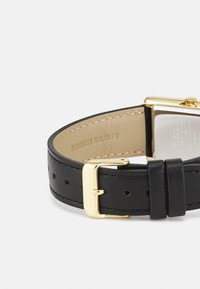 Bracelet de montre en cuir noir avec une boucle carrée en ton doré. Texture lisse avec des détails de couture et des trous de réglage visibles.