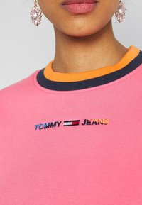 Růžová mikina s oranžovým a tmavě modrým žebrovaným límcem, zdobená vyšívanými logy "TOMMY" a "JEANS" a texturovaným povrchem látky.