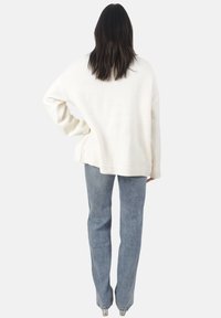 Kobieta stoi tyłem do kamery, ubrana w biały oversize'owy sweter, niebieskie dżinsy i przezroczyste botki na obcasie, na białym tle.