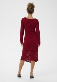 Abito rosso in pizzo con maniche lunghe, silhouette aderente e lunghezza sotto il ginocchio. Presenta un collo tondo e un orlo asimmetrico.