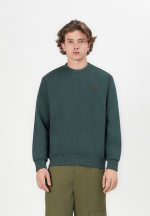 Sweatshirt vert foncé en matière douce, avec un col rond, des poignets côtelés et un petit patch logo sur la poitrine.