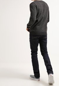 Suéter gris oscuro con motas, con dobladillo acanalado, combinado con jeans ajustados azul oscuro y zapatos grises de slip-on, visto desde la parte posterior.