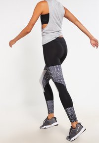 Czarne legginsy z wysokim stanem mają wzór w węże w szarych i białych akcentach. W połączeniu z szarym topem bez rękawów i butami sportowymi.