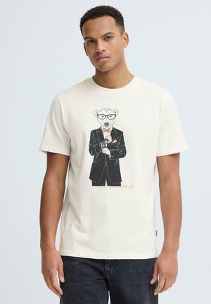 Młody mężczyzna w białej koszulce z grafiką niedźwiedzia w czarnym garniturze, okularach i muszce, poprawiający rękaw.