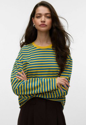 Young woman with long dark hair wearing silver hoop earrings and a yellow and blue striped long-sleeve shirt, arms crossed.