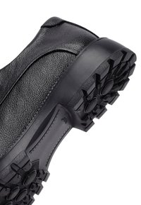 Zwarte leren schoen met een gestructureerde afwerking, voorzien van een dikke rubberen zool met onderscheidende grippatronen. Gebogen ontwerp en minimale stiksels.
