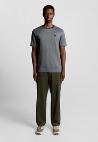Grå kortærmet T-shirt med rund halsudskæring og et diskret logo på venstre bryst, parret med olivengrønne bukser og neutrale sneakers.