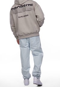 Sweat à capuche gris clair avec texte noir au dos, associé à un jean ample bleu clair et des baskets blanches. Design simple et décontracté.