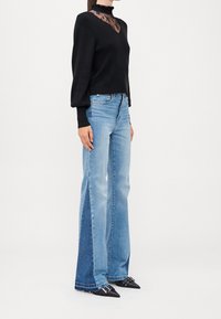 Maglione nero con scollo in pizzo, abbinato a jeans a zampa di elefante azzurri con pannelli in denim scuro a contrasto. Scarpe nere con punta appuntita.