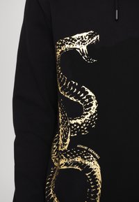 Crna kapuljača s velikim zlatnim grafikonom zmije. Materijal je glatkog završetka, s opuštenim krojem. Logo "Armani Exchange" vidljiv je u malom fontu.