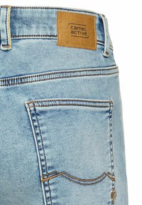 Jean en denim bleu clair avec une étiquette en cuir brun au niveau de la taille. Présente des coutures jaunes et un design de poche standard.