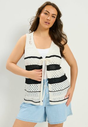 Kvinde iført ærmeløs strikket vest med sorte og hvide striber over en hvid top, kombineret med lyseblå denimshorts, poserer med den ene hånd på hoften.