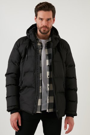 Sort puffet jakke med hætte, der har en glat tekstur og en ballonformet design. Båret over en ternet skjorte i sort og hvid, med en grå T-shirt under.
