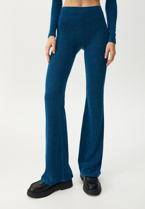 Kobieta nosząca niebieskie, szerokie spodnie z wysokim stanem oraz czarne buty na platformie, stojąca na jednolitym białym tle.