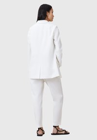 Blazer bianco su misura e pantaloni abbinati, tessuto liscio, silhouette strutturata, dettaglio della fessura posteriore, abbinati a sandali neri.