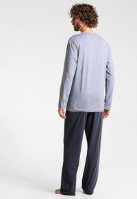 Langærmet grå top med rund halsudskæring, parret med mørkegrå loungebukser. Stoffet ser blødt ud med en afslappet pasform. Barfodet stilling.