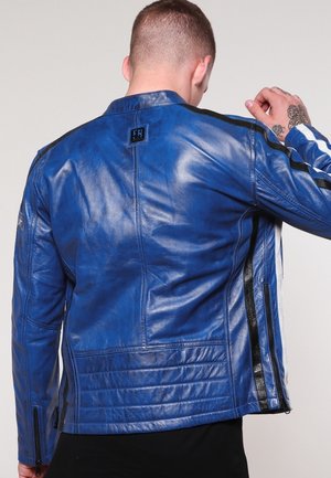 Homme portant une veste en cuir bleue brillante avec des rayures noires et blanches sur les manches, de dos, la main droite levée près de la tête.