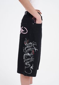 Pantalones cortos de mezclilla negra con un intrincado bordado de dragón en gris y rojo. Presenta un gráfico rosa en la parte posterior y bolsillos con forro floral.