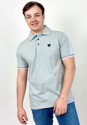 Jeune homme souriant, portant un polo gris clair avec des boutons foncés et un jean bleu, debout sur un fond blanc.