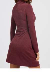 Femme portant une robe ajustée, à manches longues, en côte bordeaux avec un col roulé, vue de dos sur un fond uni.