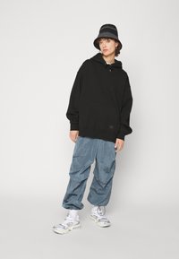 Sudadera negra de gran tamaño con un bolsillo frontal y cordón, combinada con pantalones cargo de tela texturizada en un azul claro y zapatillas gruesas blancas.