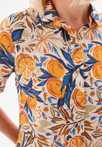 Short-sleeve shirt in lightweight fabric with a vibrant floral pattern in orange, blue, and green on a cream background. Button-up design.