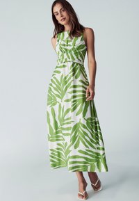 Maxi haljina s oblikovanim korzetom i lepršavom suknjom, izrađena od bijelog materijala s velikim zelenim uzorcima lišća. Bijele sandale s trakama upotpunjuju izgled.