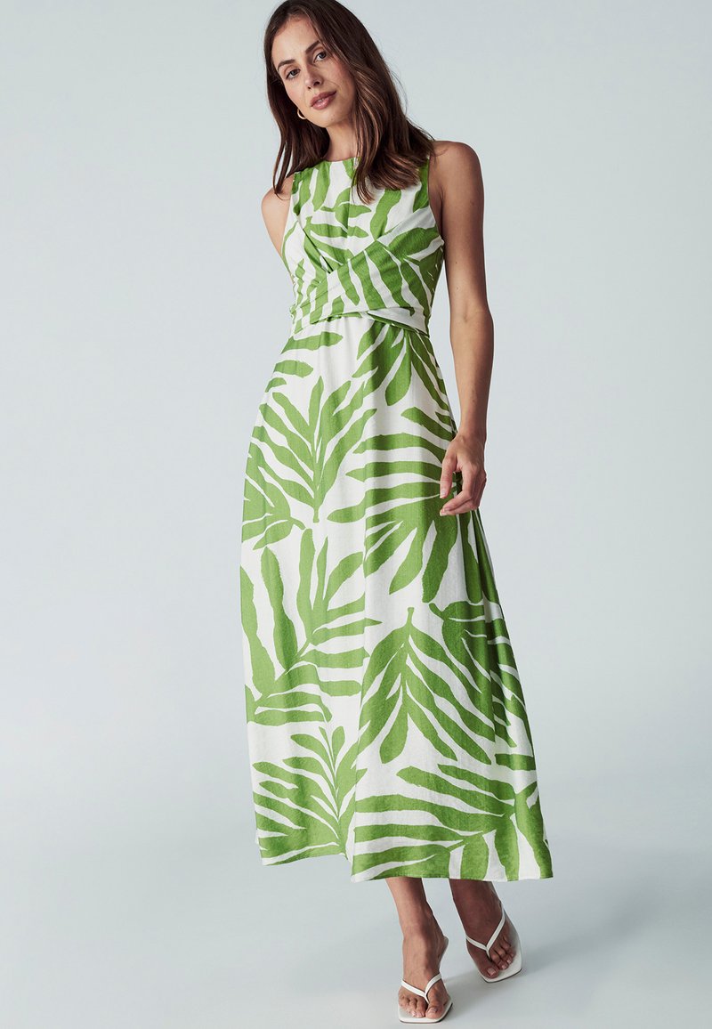 Maxi haljina s oblikovanim korzetom i lepršavom suknjom, izrađena od bijelog materijala s velikim zelenim uzorcima lišća. Bijele sandale s trakama upotpunjuju izgled.