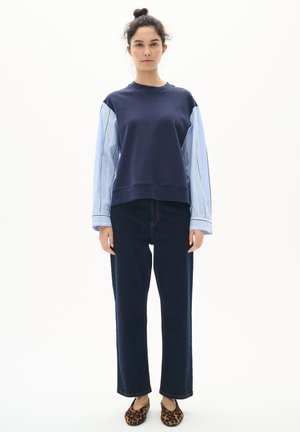 Marineblauer Sweatshirt mit hellblauen gestreiften Ärmeln, kombiniert mit dunkelblauen Jeans im Straight-Leg-Schnitt und flachen Schuhen im Leopardenmuster. Schlichtes, modernes Design.