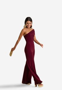 Combinaison bordeaux, décolletée sur les épaules, avec un corsage ajusté et un pantalon à jambes larges en tissu doux et extensible. Les accessoires comprennent une pochette.