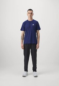 Tamsiai mėlynas sportinis marškinėlis su tekstūruotu raštu ir baltu logotipu, derinamas su juodomis kelnėmis ir baltais sportbačiais, dėvimas stovinčio vyro modelio.