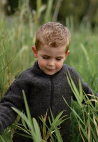 Barn som bär en mörkgrå fleecetröja med hög krage och dragkedja fram, omgiven av hög grön gräs. Håret är ljust och kort.