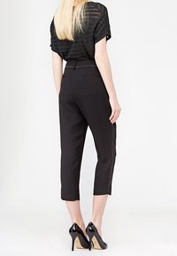 Černé zkrácené kalhoty s elegantním, na míru šitým střihem, s hladkou texturou a bočními kapsami. Doplněné lesklými černými vysokými podpatky.