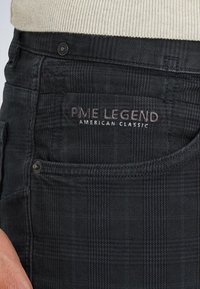 Närbild av en ficka på svarta rutiga byxor med PME Legend American Classic broderat ovanför fickan och metallnitar på sömmarna.