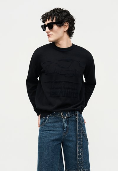 Άνδρας με σγουρά μαλλιά που φοράει μαύρα γυαλιά ηλίου, μαύρο φούτερ με σχέδιο Snoopy και μπλε τζιν τζιν με ζώνη με κρίκους.