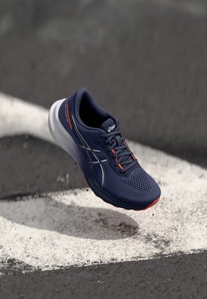 Marineblauer Laufschuh mit orangen Akzenten und weißer Sohle, schwebend über Asphalt mit weißer geschwungener Markierung.