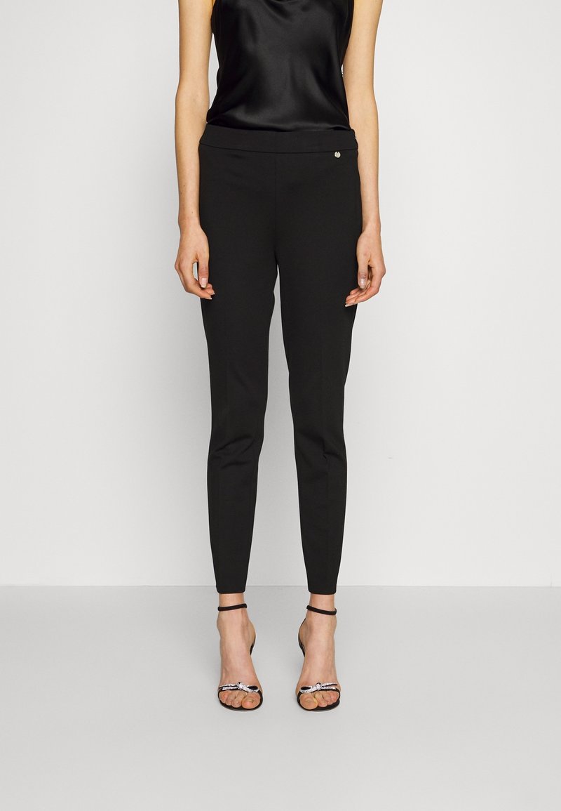 Pantalon noir ajusté en tissu lisse, coupe slim, longueur cheville, avec un petit accent métallique à la taille. Associé à un haut en soie et des talons.