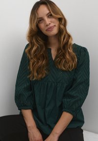 Temno zelena bluza z teksturirano, chevron vzorcem, z balonastimi rokavi in V-izrezom. Narejena iz mehke, zračne tkanine.