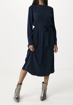 Woman wearing a long-sleeve navy midi dress with a waist tie and metallic silver ankle boots, standing against a plain background.