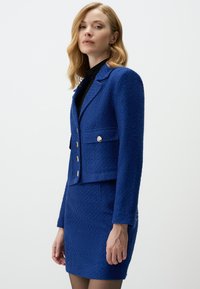 Blue textured jacket with a collar, front pockets, and silver buttons, paired with a matching fitted skirt.