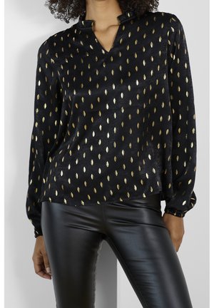 Schwarze langärmlige Bluse mit goldenen Tropfenmustern, kombiniert mit schwarzen Hosen aus Kunstleder, getragen von einem Model von Hals bis Oberschenkel.