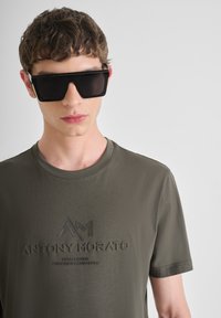 Camiseta de manga corta verde oliva con el logotipo "ANTONY MORATO" en relieve, combinada con gafas de sol negras angulares. Textura suave, diseño moderno.